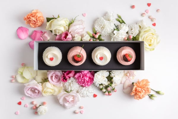 Box of cupcakes decorated with strawberries and heart sprinkles, surrounded by flowers