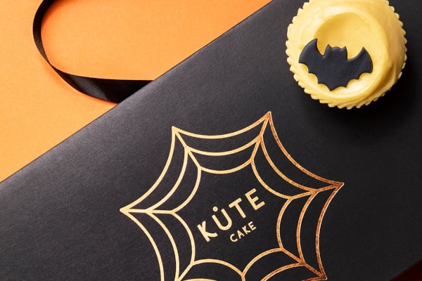 Black box with Kute Cake logo inside spider web and yellow cupcake with bat on top