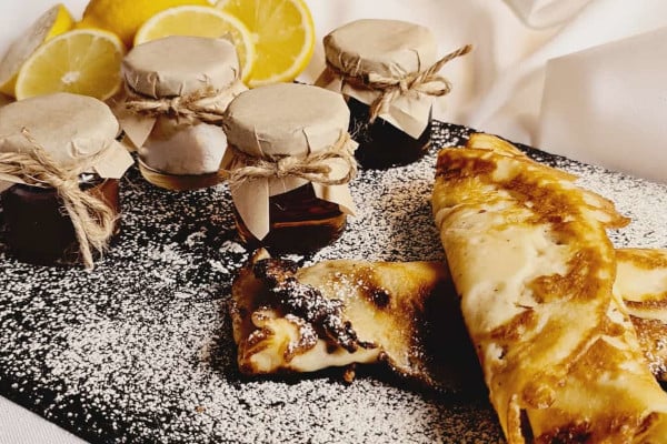 Rolled up pancakes on slate board with dusting of icing sugar next to pots of jam and lemon slices